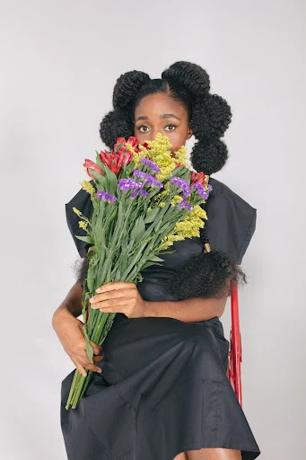 Attractive Black woman with styled natural hair poses holding a colorful bouquet in a studio setting, wearing a stylish black dress.