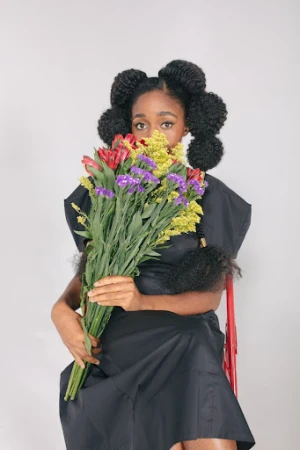 Attractive Black woman with styled natural hair poses holding a colorful bouquet in a studio setting, wearing a stylish black dress.