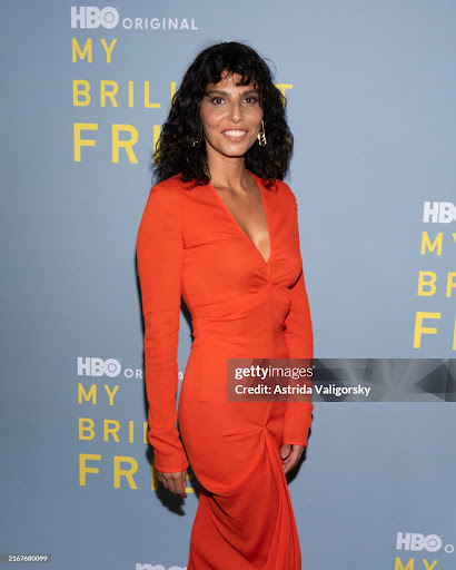 Portrait of attractive adult woman in orange dress standing at event for 'My Brilliant Friend', smiling at the camera, indoors.