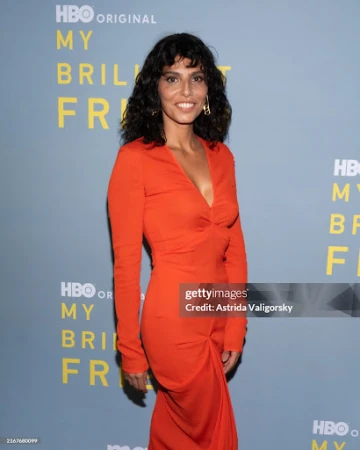 Portrait of attractive adult woman in orange dress standing at event for 'My Brilliant Friend', smiling at the camera, indoors.