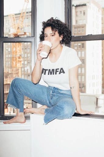 Young woman with curly hair sits on a window ledge in jeans and a white t-shirt drinking coffee with a cityscape background.