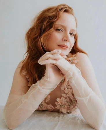 Portrait of a red-haired woman wearing sheer gloves and a floral embellished gown against a neutral background.