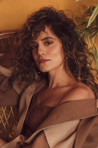 Brunette woman with wavy hair poses indoors wearing a camel-colored coat and brown dress in a warm-toned, soft focus portrait.