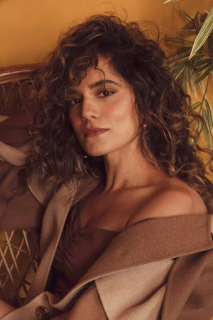 Brunette woman with wavy hair poses indoors wearing a camel-colored coat and brown dress in a warm-toned, soft focus portrait.