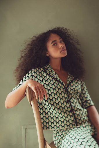 Young woman with curly hair sits in a patterned shirt and skirt, posing indoors in front of a solid light green wall.