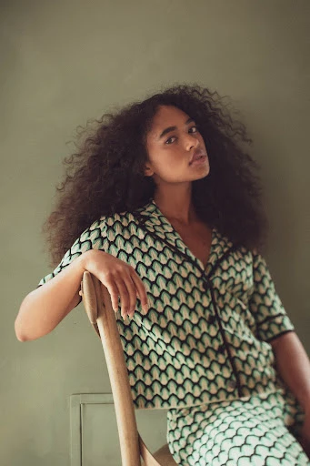 Young woman with curly hair sits in a patterned shirt and skirt, posing indoors in front of a solid light green wall.
