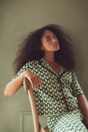 Young woman with curly hair sits in a patterned shirt and skirt, posing indoors in front of a solid light green wall.