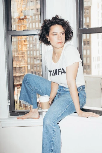 Adult female sitting on a window sill in jeans and a t-shirt holding a cup with a city building visible in the background.