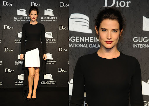 Cobie Smulders poses in a black long-sleeve dress with a white panel at an event with a black backdrop featuring brand logos and text.
