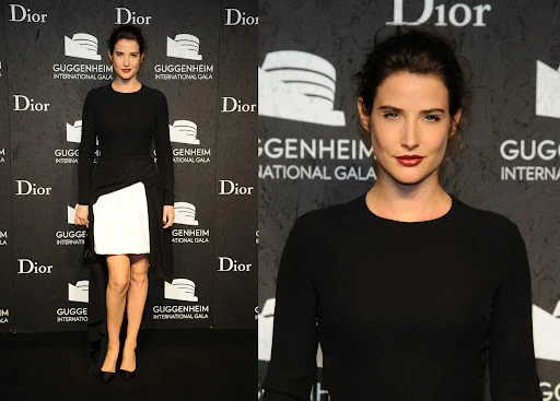 Cobie Smulders poses in a black long-sleeve dress with a white panel at an event with a black backdrop featuring brand logos and text.