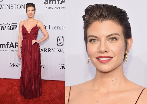 Lauren Cohan poses in a long burgundy gown on a red carpet with various brand logos and text visible in the background.