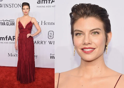 Lauren Cohan poses in a long burgundy gown on a red carpet with various brand logos and text visible in the background.