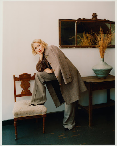 Elegant woman in a pantsuit poses indoors. She sits on a wooden chair with a vase of dried flowers and a mirror nearby.