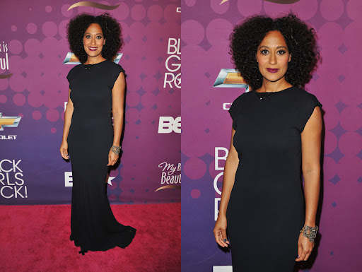 Tracee Ellis Ross poses on the red carpet in a black gown with her hair styled in a large afro. The event backdrop is purple.