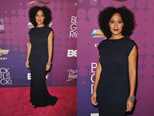 Tracee Ellis Ross poses on the red carpet in a black gown with her hair styled in a large afro. The event backdrop is purple.
