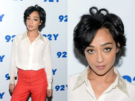 Portrait of actress Ruth Negga wearing a white collared blouse and orange slacks in front of a gray backdrop.