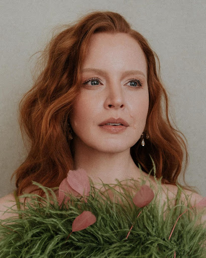 Close-up of a red-haired woman with fair skin, adorned with a decorative green and pink feathered collar, indoors.