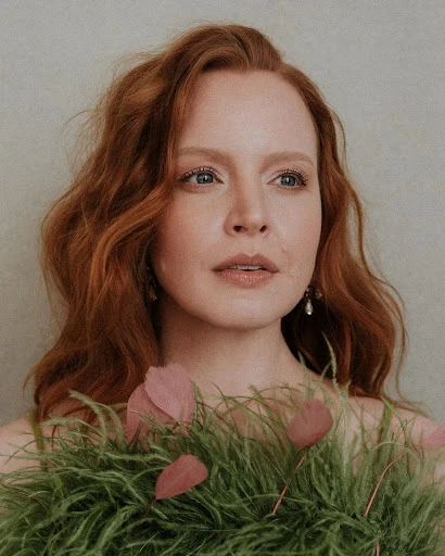 Close-up of a red-haired woman with fair skin, adorned with a decorative green and pink feathered collar, indoors.