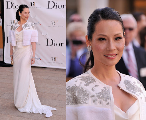 Lucy Liu on the red carpet in an off-white gown with a white faux fur bolero with silver embroidery, Dior signage backdrop. 