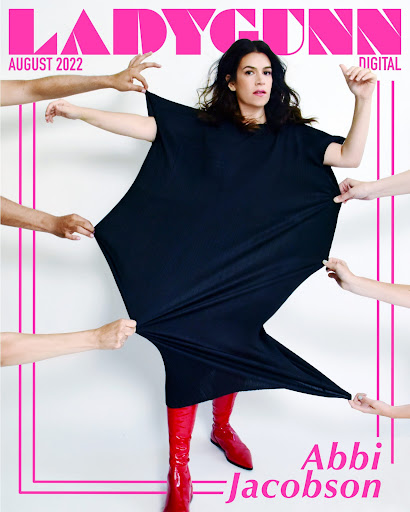 Magazine cover featuring Abbi Jacobson wearing a black dress and red boots, stretched by surrounding arms, against a white backdrop.