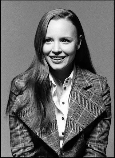 Portrait of a smiling woman with long hair wearing a plaid blazer and button-down shirt against a textured grey background.