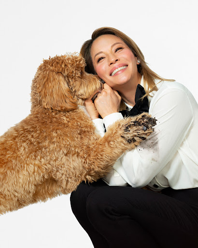 Adult woman in a white button-down shirt and black pants is posing with a tan-colored Goldendoodle dog in a studio setting.