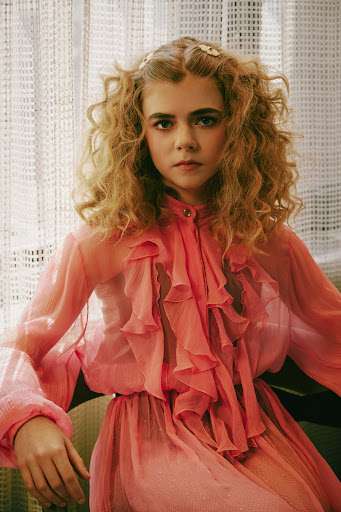 Portrait of a blonde woman with curly hair, wearing a pink dress with ruffles, sitting in a chair with a patterned curtain background.