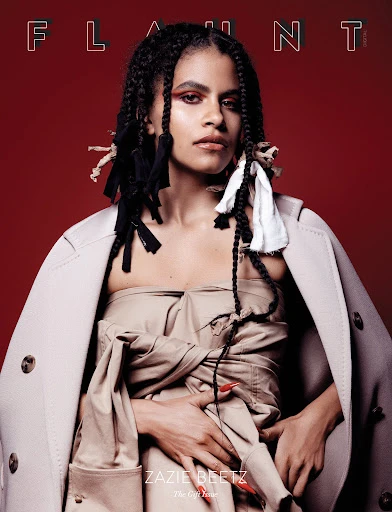 Zazie Beetz poses for a fashion magazine cover with a red background, wearing a beige jacket and strapless top with braided hair.