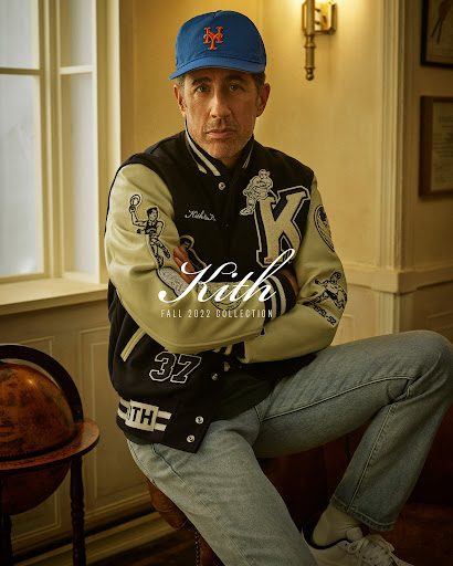 Jerry Seinfeld wearing a Kith letterman jacket and blue baseball cap seated indoors with arms crossed