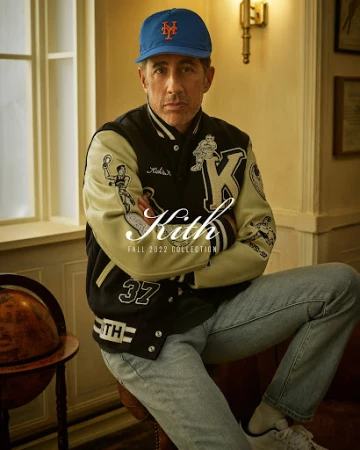 Jerry Seinfeld wearing a Kith letterman jacket and blue baseball cap seated indoors with arms crossed