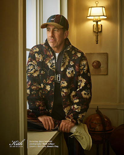 Jerry Seinfeld poses in a floral jacket and cap inside a building with warm lighting, creating a stylish and inviting scene.