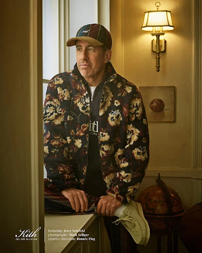Jerry Seinfeld poses in a floral jacket and cap inside a building with warm lighting, creating a stylish and inviting scene.