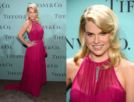 Alice Eve poses in a full-length, magenta chiffon gown on a Tiffany & Co. branded backdrop, captured in an indoor setting with flattering lighting.