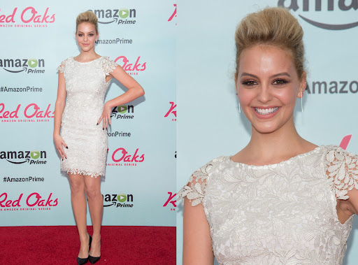 A woman in a white lace dress and black heels poses on a red carpet at an Amazon Prime Red Oaks event, smiling and looking at the camera.