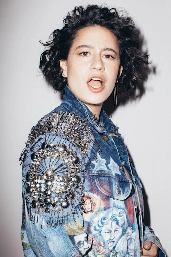 Young woman in embellished denim jacket posing indoors against a neutral backdrop, portrait shot, eye-level view, studio lighting.