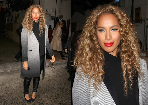 Stylish woman with blonde curly hair wears a black top, gray coat, black pants and heels, standing outdoors in an urban setting.