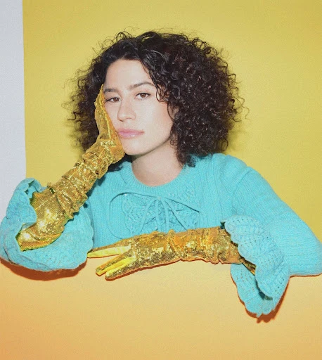 Portrait of a young woman with curly hair and golden gloves, wearing a blue sweater, posing against a yellow and white background.