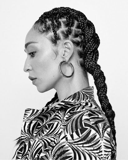 Portrait of an adult woman in profile wearing patterned shirt and large hoop earring, with braided hair against a white background.