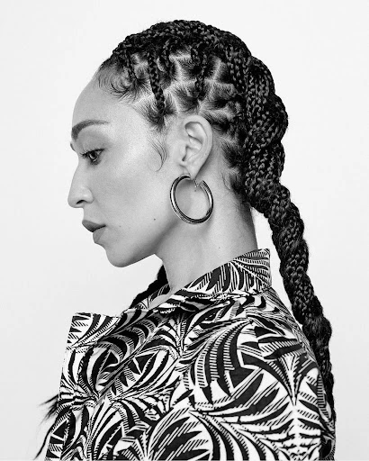 Portrait of an adult woman in profile wearing patterned shirt and large hoop earring, with braided hair against a white background.
