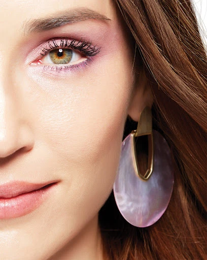 Close-up studio shot of a fair-skinned adult woman with hazel eyes and pink eye shadow, wearing gold and lavender teardrop earrings.