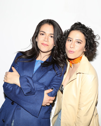 Two women posing together against a white wall, one wearing a blue blazer and the other in a beige jacket with an orange neck scarf.