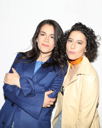 Two women posing together against a white wall, one wearing a blue blazer and the other in a beige jacket with an orange neck scarf.