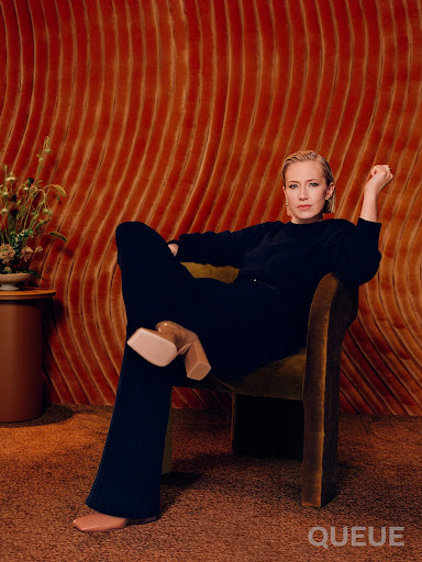 Adult woman in a black outfit sits in a green chair with a patterned, textured orange wall in the background and a flower arrangement.