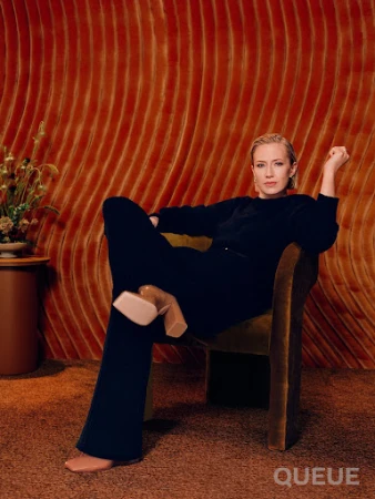 Adult woman in a black outfit sits in a green chair with a patterned, textured orange wall in the background and a flower arrangement.
