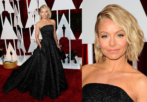 Kelly Ripa at a red carpet event in a black strapless gown, posing in front of a backdrop, her hair blonde and wavy.