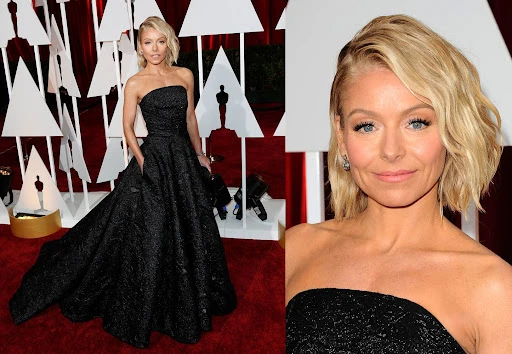 Kelly Ripa at a red carpet event in a black strapless gown, posing in front of a backdrop, her hair blonde and wavy.