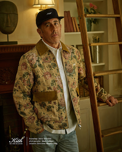 Portrait of Jerry Seinfeld wearing a floral jacket and a baseball cap, standing next to a wooden ladder indoors.