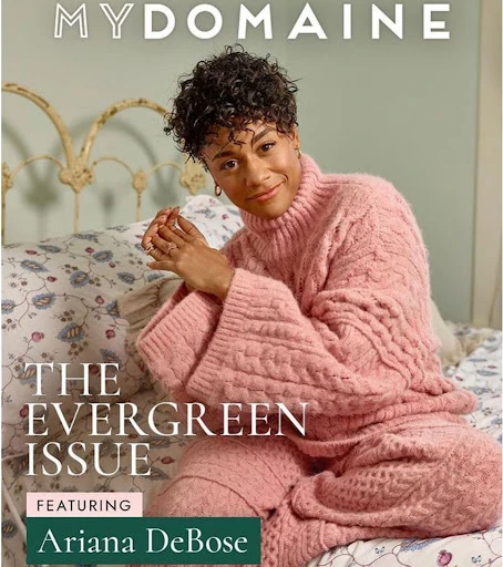 Ariana DeBose poses for the cover of MYDOMAINE's The Evergreen Issue, wearing a pink sweater and matching pants on a bed.