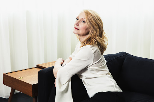 Portrait of a woman with blonde hair sitting on a blue couch, wearing a cream-colored blouse and black pants, with white curtains in the background.