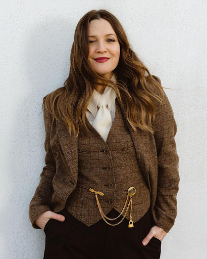 Drew Barrymore wearing a brown tweed blazer, vest, and dark pants, posing against a white wall in a medium studio shot.